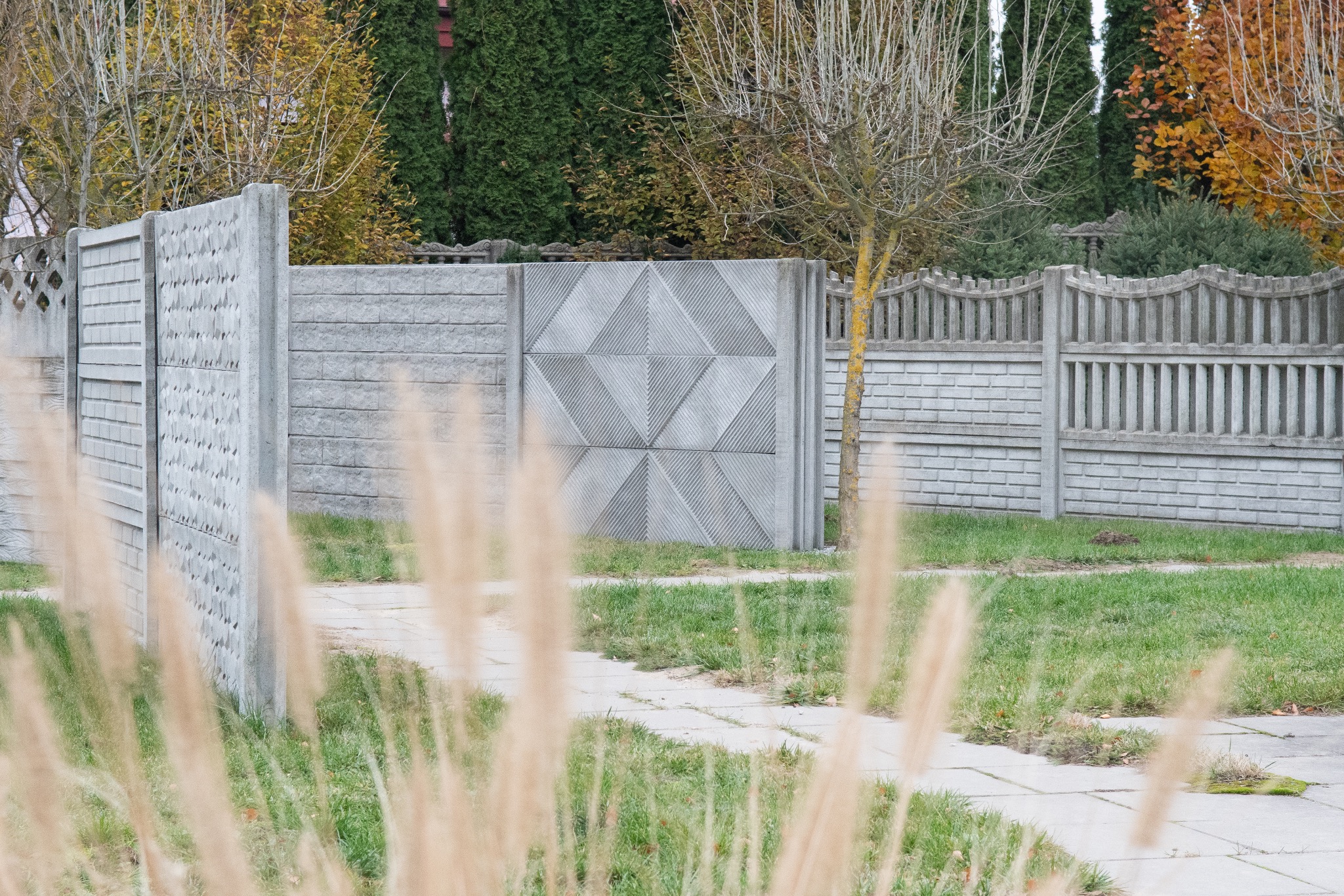 Fragmenty betonowego ogrodzenia o różnych wzorach, widoczne przez źdźbła trawy, na tle zieleni i drzew.
