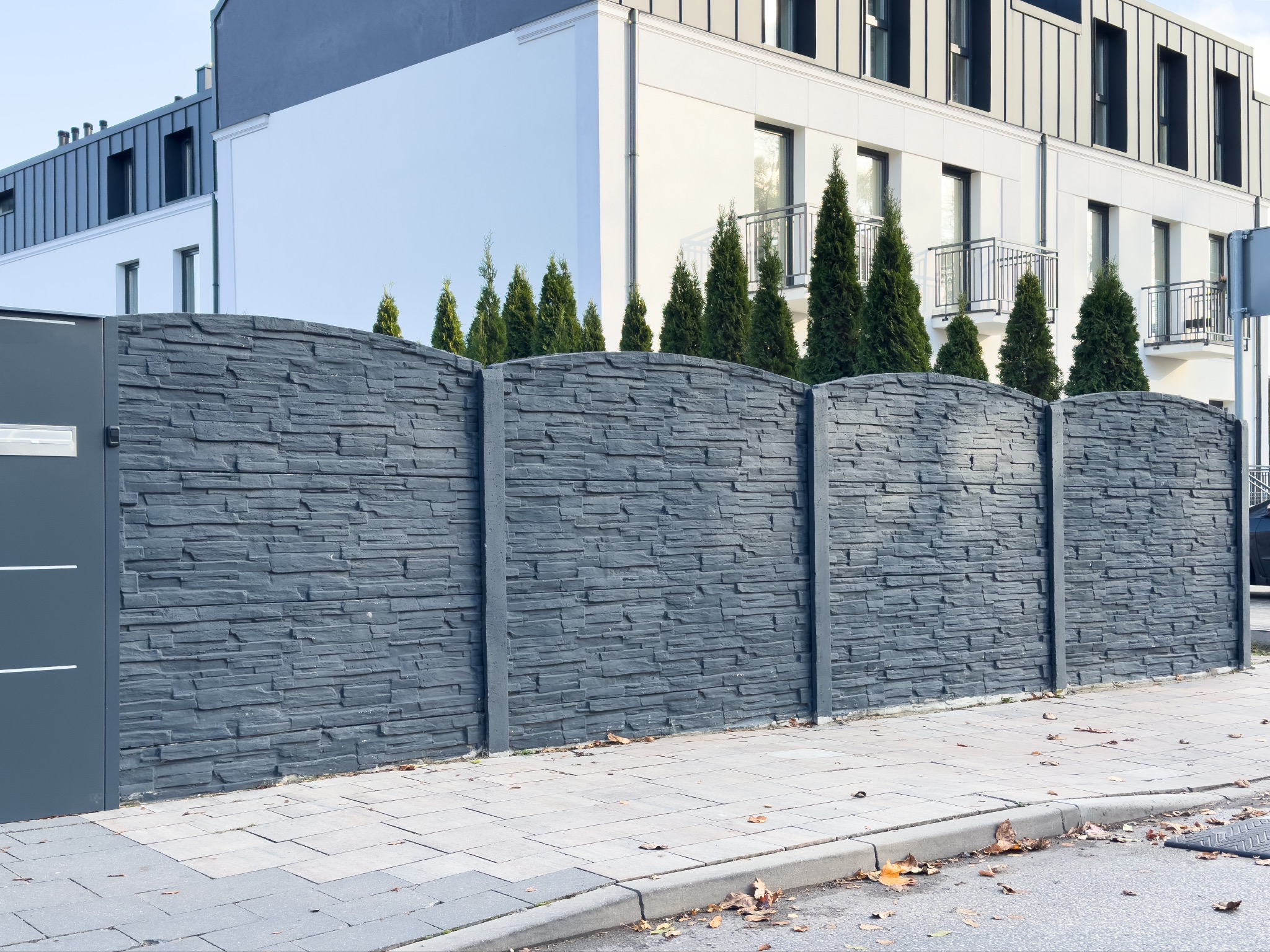Nowoczesne, szare ogrodzenie z betonu imitującego kamień, z przesuwną bramą w kolorze antracytowym, na tle białego budynku z ciemnymi oknami i rzędem małych drzew iglastych.