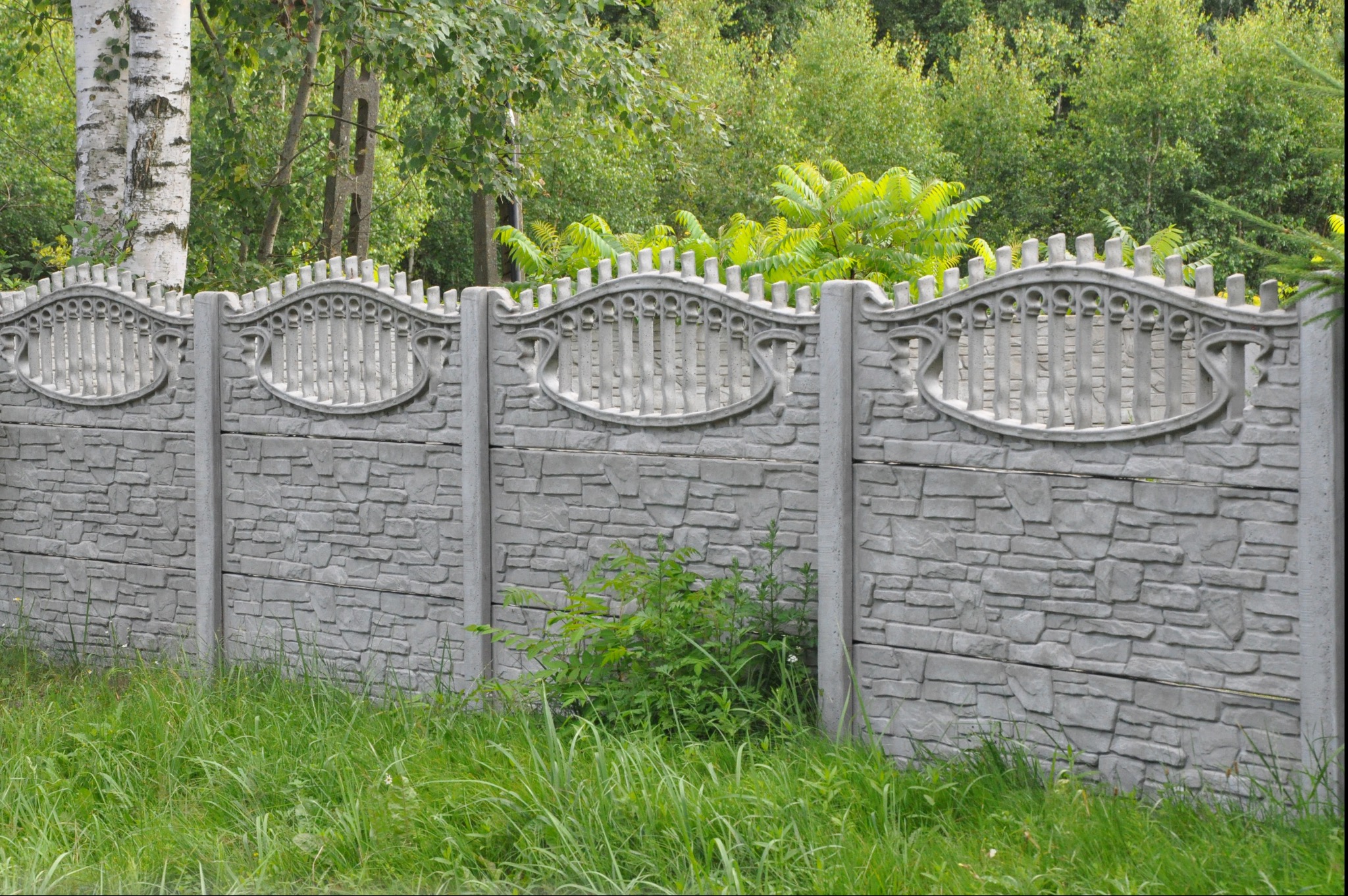 Szary, prefabrykowany płot betonowy z dekoracyjnymi ażurowymi wstawkami, widok z poziomu gruntu na tle zieleni.