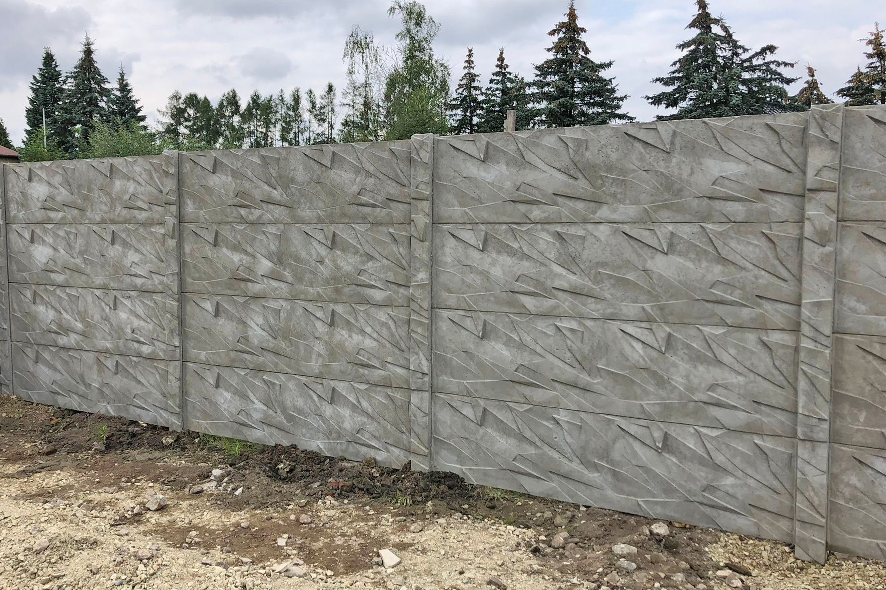 Szary, prefabrykowany płot z betonu z abstrakcyjnym wzorem, widok na drzewa w tle.