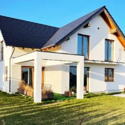 Nowoczesny dom jednorodzinny z białymi ścianami, ciemną dachówką i drewnianymi oknami, widziany z perspektywy ogrodu w słoneczny dzień.