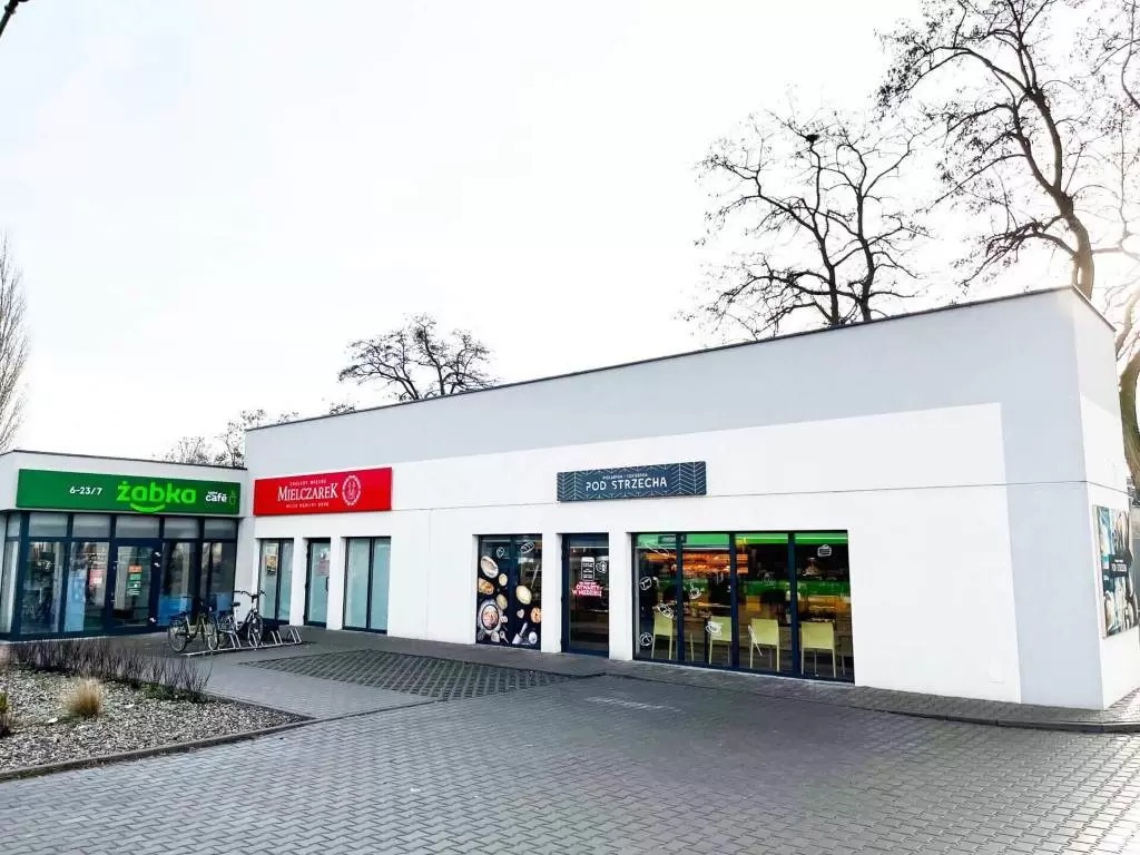 Parterowy budynek handlowy z płaskim dachem, mieszczący lokale Żabka, Mielczarek i Pod Strzechą, z rowerami zaparkowanymi przed wejściem do Żabki.