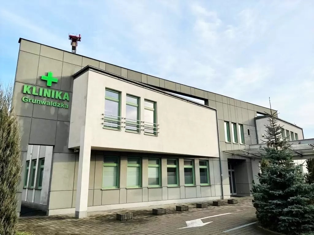 Nowoczesny budynek kliniki z szarymi panelami i białymi elementami architektonicznymi, zielonym logo i oknami, widoczny z zewnątrz, z kostką brukową i strzałką na parkingu.