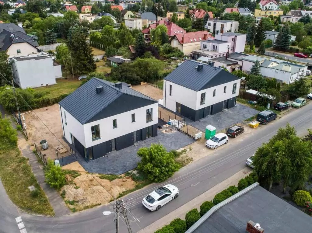 Dwa nowoczesne domy murowane z szarą podmurówką i ciemnoszarym dachem, widok z góry, teren wokół w trakcie zagospodarowywania, na ulicy zaparkowane samochody i przejeżdżający biały samochód...