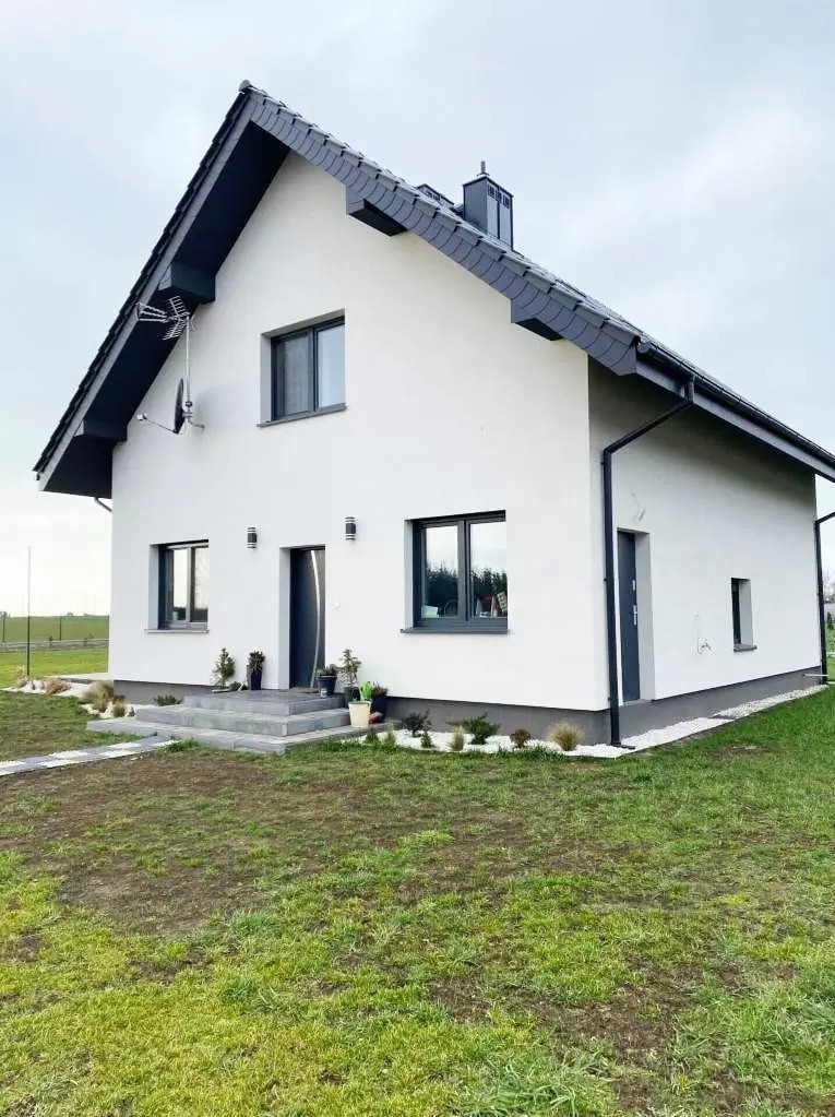 Nowoczesny, dwukondygnacyjny dom z białymi ścianami, ciemnoszarą dachówką i stolarką okienną, z anteną satelitarną na elewacji, otoczony trawnikiem z młodą trawą.