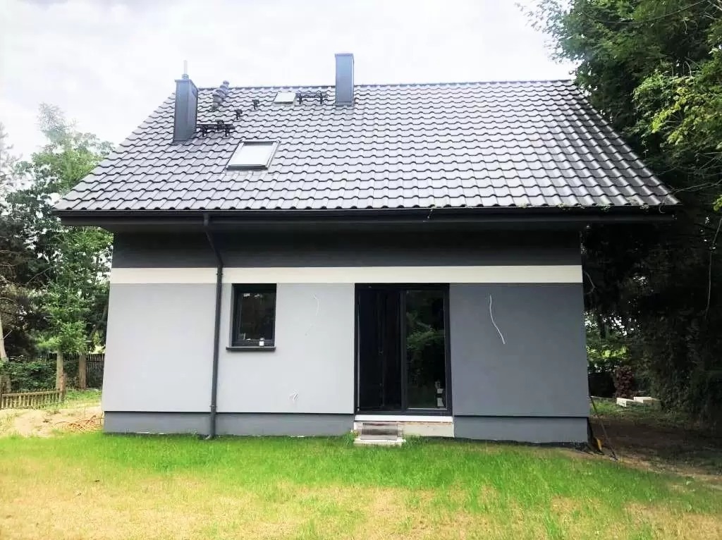 Nowoczesny, jednopiętrowy dom z szarą elewacją, ciemną dachówką i dwoma kominami, widoczny z poziomu trawnika.
