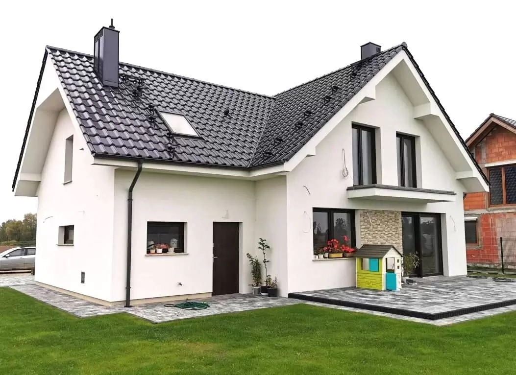 Nowoczesny, biały dom jednorodzinny z czarnym dachem i oknami, z tarasem wyłożonym szarą kostką, na którym stoi mały, kolorowy domek dla dzieci.