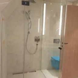 Nowoczesna kabina prysznicowa z marmurowymi płytkami, szklanymi drzwiami i chromowaną armaturą. Uwagę zwraca ukryty odpływ liniowy w podłodze i toaleta zabezpieczona folią ochronną.