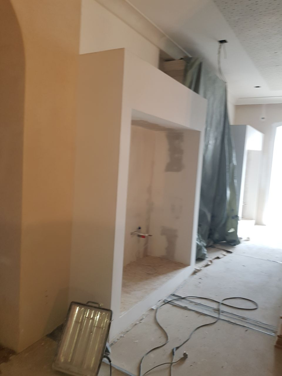 Wnętrze w trakcie prac wykończeniowych: wnęka w ścianie przygotowana do zabudowy, ściany w surowym stanie, podłoga z widocznymi śladami prac, reflektor halogenowy na podłodze, przewody elektryczne...