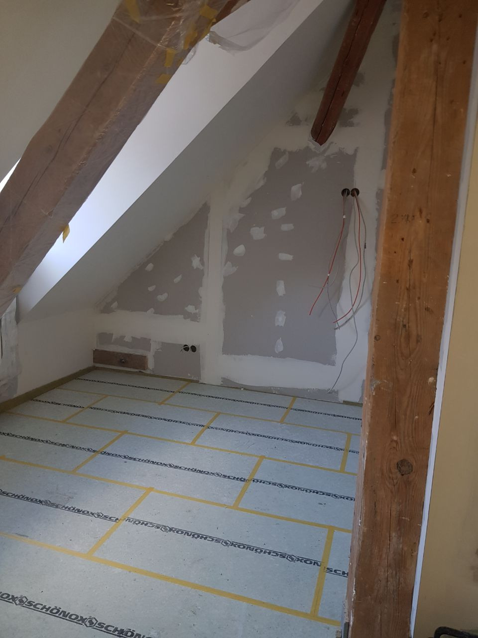 Poddasze w trakcie prac wykończeniowych: widoczne drewniane belki, ściany przygotowane pod gładzie, zaklejone taśmą płyty podłogowe, wystające przewody elektryczne.