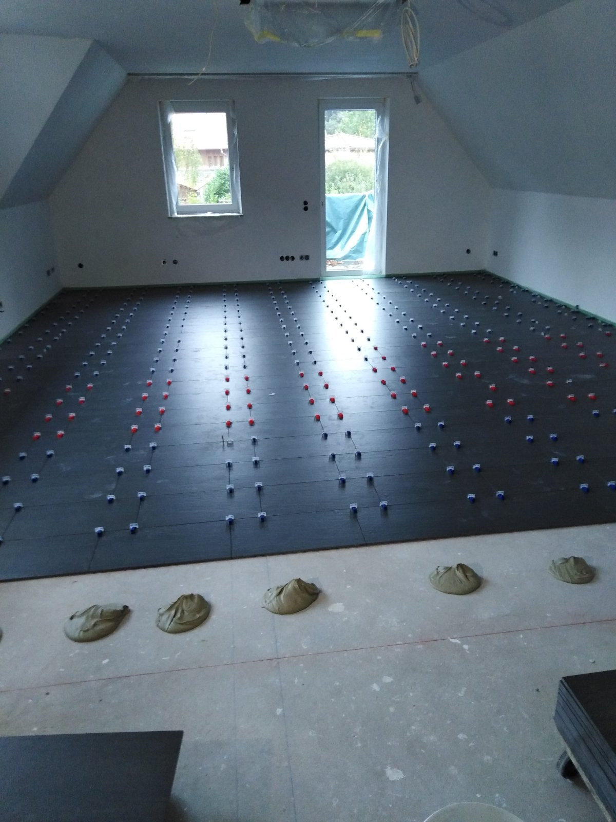 Montaż ciemnych płytek podłogowych w pomieszczeniu na poddaszu, widoczne krzyżyki dystansowe i porcje kleju na surowej posadzce.