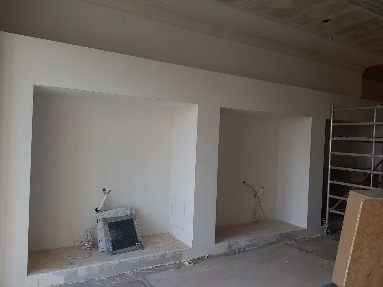 Dwa wnęki w ścianie po świeżym malowaniu na biało, widoczne wystające kable instalacji elektrycznej, na podłodze resztki gruzu i narzędzia, w tle rusztowanie.