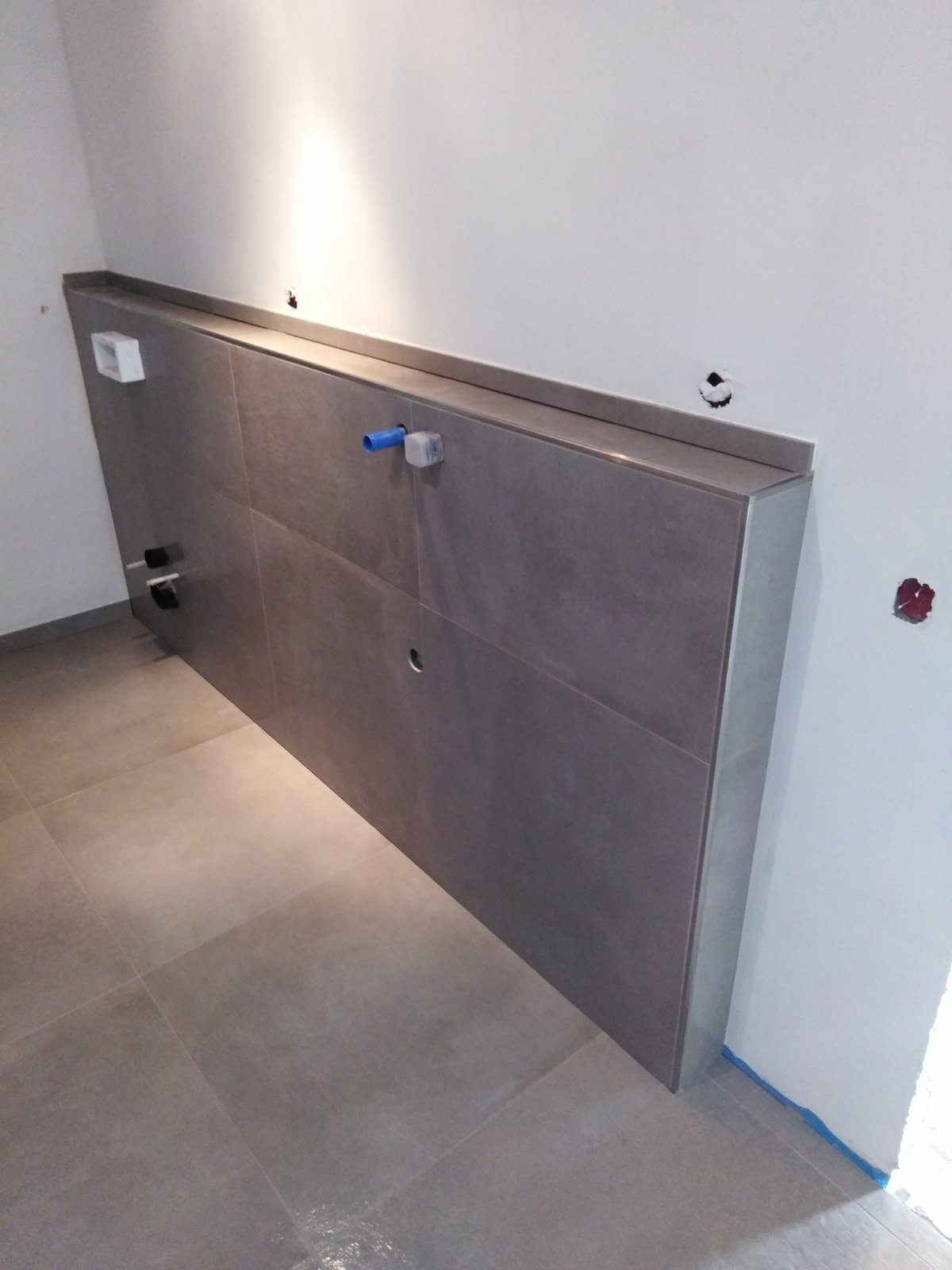 Szara zabudowa stelaża WC z płytek ceramicznych w trakcie montażu, widoczne wystające rury i otwory w ścianie, podłoga wyłożona płytkami w kolorze szarym.