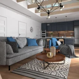 Jasne wnętrze domu z szarym narożnikiem, drewnianym stołem kawowym i schodami drabinkowymi, z widokiem na otwartą kuchnię z ciemnymi szafkami i drewnianym blatem.