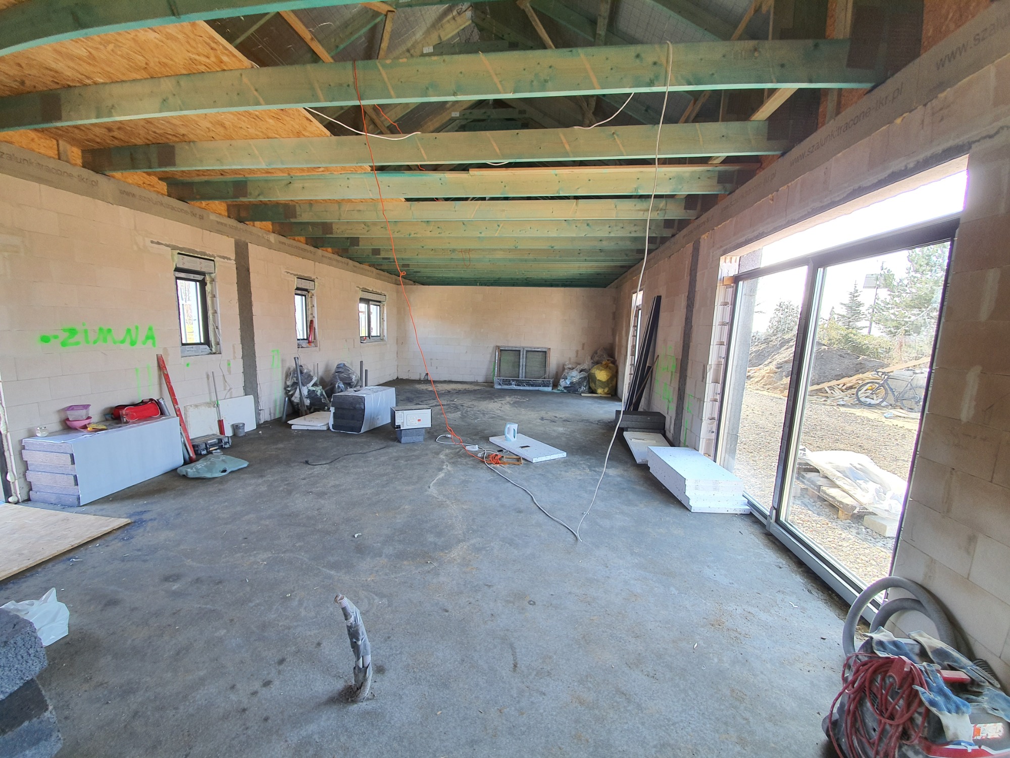 Wnętrze surowego domu murowanego w trakcie budowy, widoczne bloczki, drewniana konstrukcja dachu, okna, materiały budowlane i narzędzia.