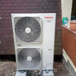 Zewnętrzna jednostka pompy ciepła Toshiba Estia, zamontowana na betonowych bloczkach na tle ściany z dachówki bitumicznej.