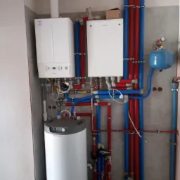 Kocioł grzewczy, zasobnik ciepłej wody użytkowej i instalacja z rur w kolorach czerwonym i niebieskim, zamontowane na tle surowej ściany w trakcie wykańczania.
