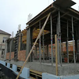 Wznoszenie budynku żelbetowego z widocznymi szalunkami, podporami stalowymi i fragmentami muru z cegły ceramicznej.