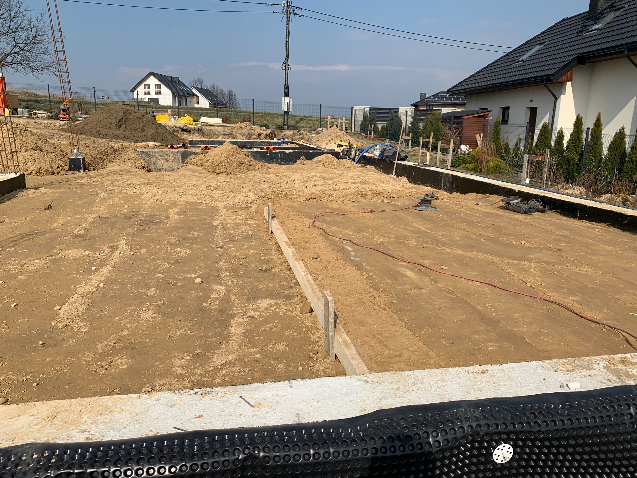 Fundamenty domu w trakcie budowy, widoczna izolacja pionowa, szalunki i wykop. W tle domy jednorodzinne i błękitne niebo. Jasne światło słoneczne.