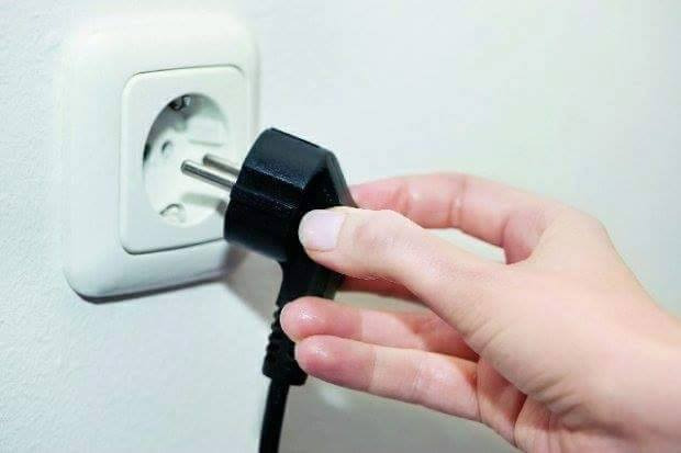 Czarny wtyk elektryczny w dłoni, zbliżający się do białego gniazdka elektrycznego na jasnej ścianie.