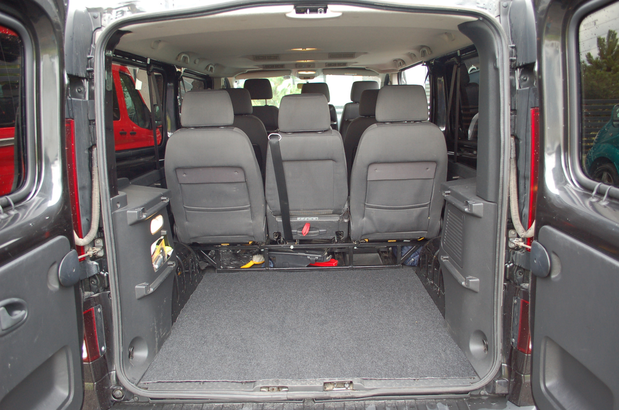 Wnętrze busa z trzema rzędami szarych foteli, widok od tyłu z otwartymi drzwiami bagażnika, szara wykładzina podłogowa.