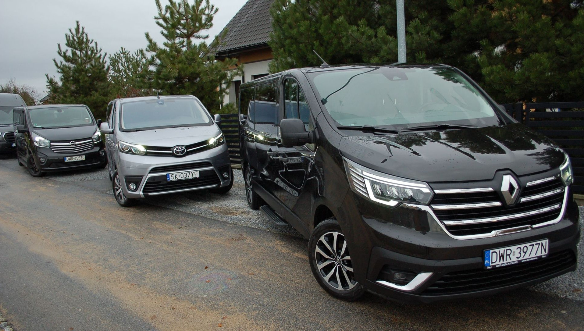 Flota samochodów osobowych: ciemnoszary Renault Trafic z widoczną tablicą rejestracyjną DWR 3977N, srebrna Toyota Proace z tablicą SK 037YF oraz ciemny Opel Vivaro, zaparkowane przed domem.