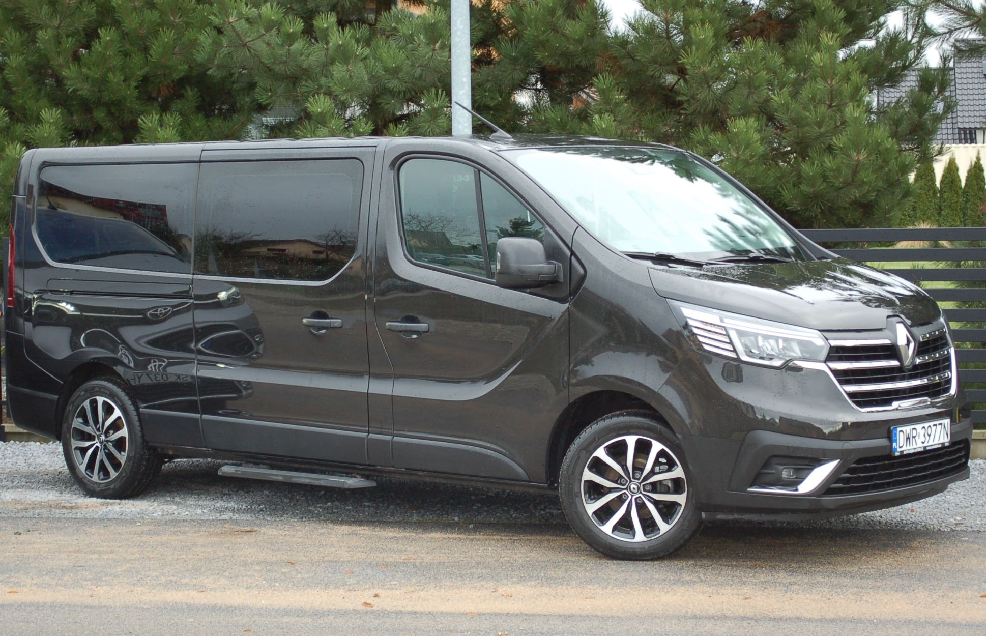 Ciemny, lśniący van Renault Trafic z aluminiowymi felgami, zaparkowany na ulicy przed zielonymi krzewami i ogrodzeniem.