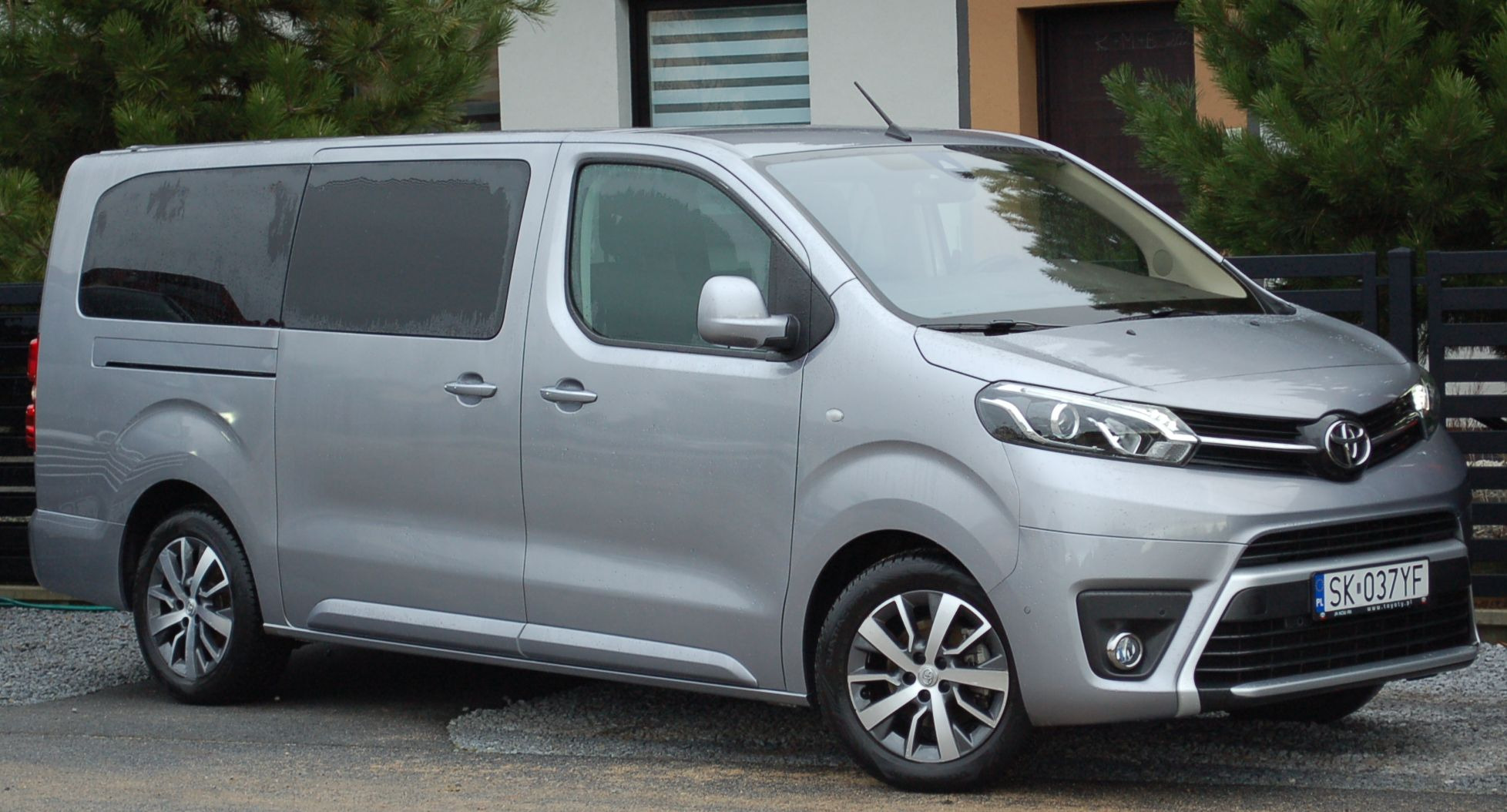 Srebrny van Toyota Proace Verso z polską tablicą rejestracyjną SK 037YF zaparkowany na żwirowym podjeździe przed budynkiem z ciemnymi oknami i zielonymi krzewami.