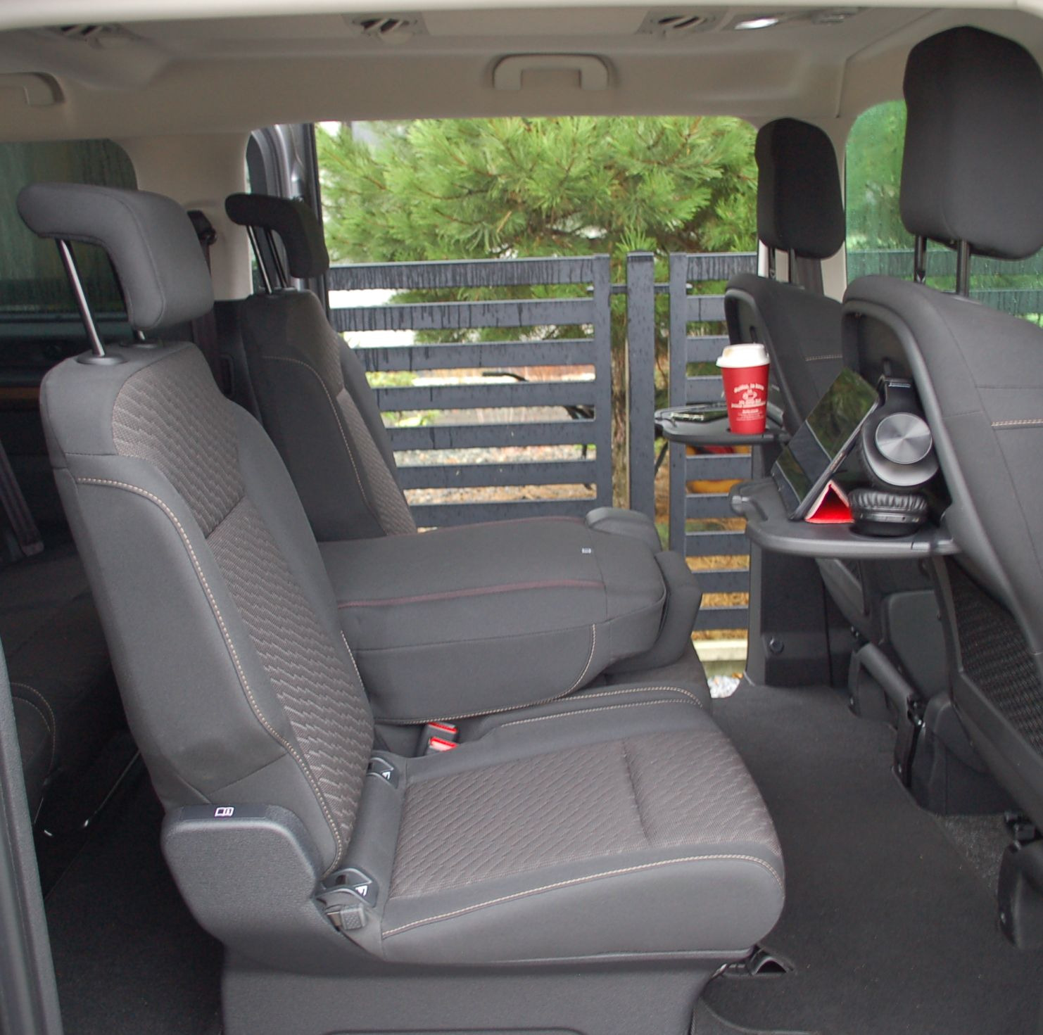 Wnętrze przestronnego vana z rozkładanymi stolikami, na których leżą tablet, słuchawki i kubek z kawą; widok z tylnych siedzeń na zewnątrz przez tylną szybę.