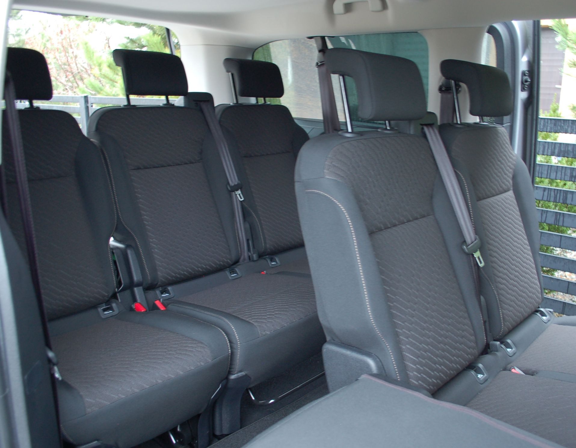 Wnętrze busa z trzema rzędami czarnych foteli z zagłówkami i pasami bezpieczeństwa, widok od strony otwartych drzwi.