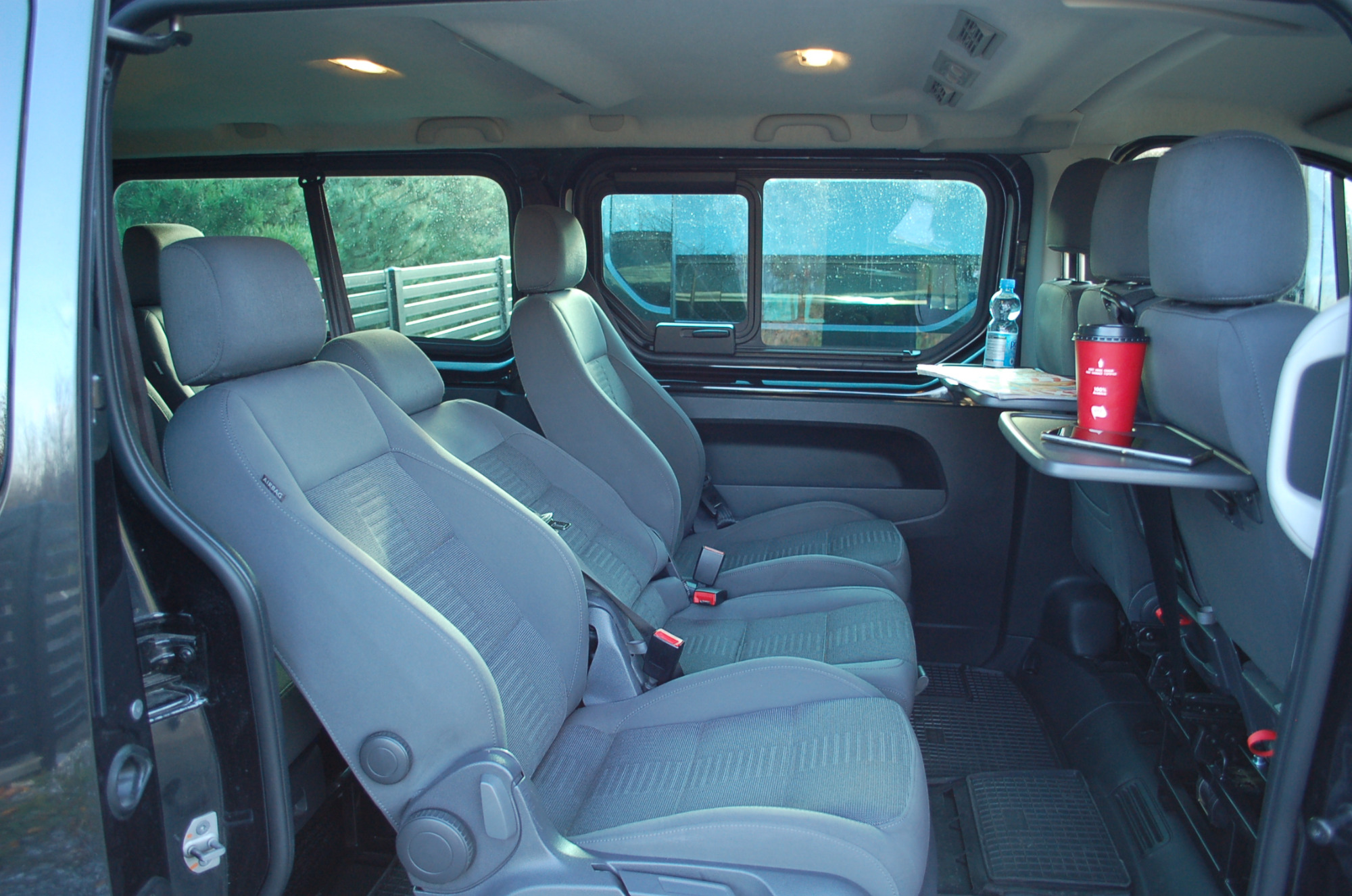 Wnętrze busa z trzema rzędami szarych foteli, stolikiem z napojem i tabletem, widok przez okno na zewnątrz.