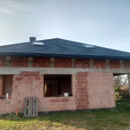 Niedokończony dom z ciemnoszarą dachówką i oknami dachowymi, ściany z czerwonej cegły, widoczna paleta i narzędzia budowlane w otoczeniu trawy.