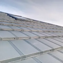 Fragment dachu pokrytego panelami na rąbek stojący w kolorze grafitowym, widoczne mocowania paneli, w tle fragment nieba.