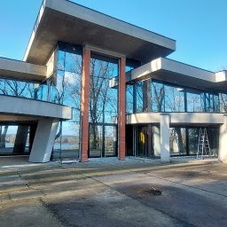 ND SYSTEM - Nowoczesny budynek z dużymi przeszkleniami, częściowo wykończony, z widocznymi elementami konstrukcyjnymi i odbiciem drzew w szybach. Na zewnątrz drabina.