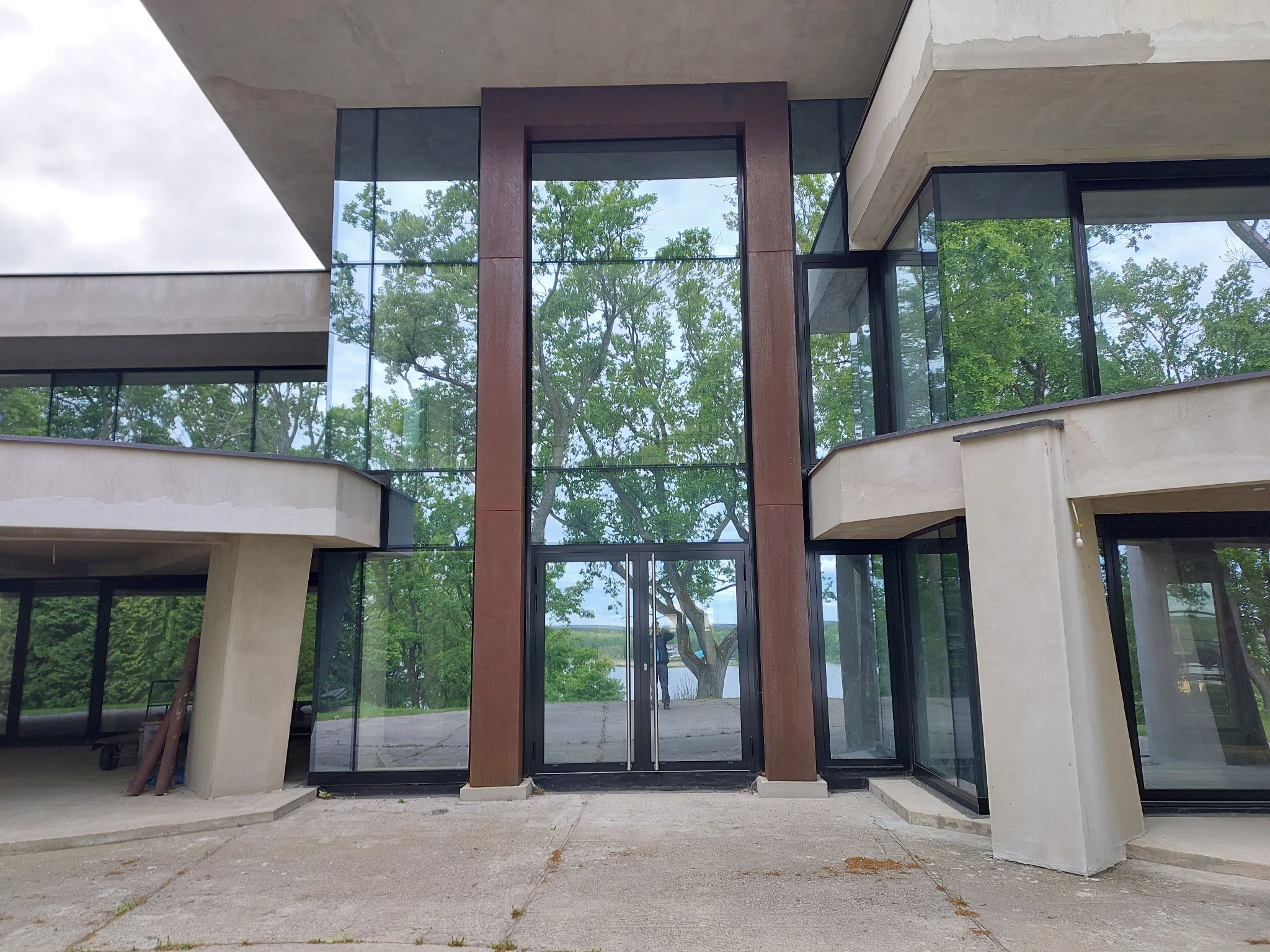 Nowoczesna fasada budynku z dużymi przeszkleniami i ciemnymi ramami okiennymi. W oknach odbija się zieleń drzew i błękit nieba, tworząc harmonijny widok.