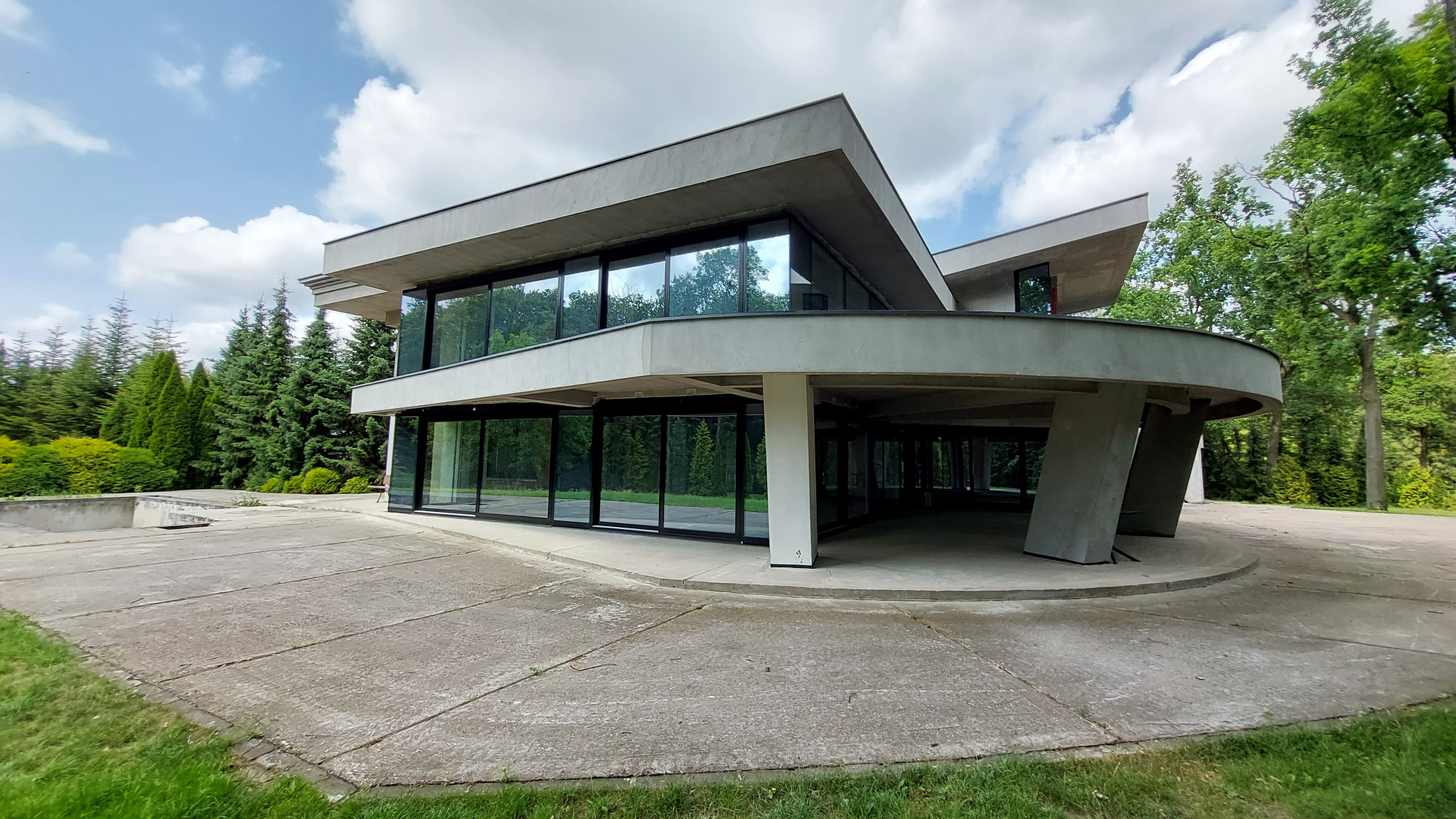 Nowoczesny dom z aluminiowymi oknami, panoramiczne przeszklenia odbijają zieleń drzew. Minimalistyczna bryła budynku, surowy beton i duże okna.