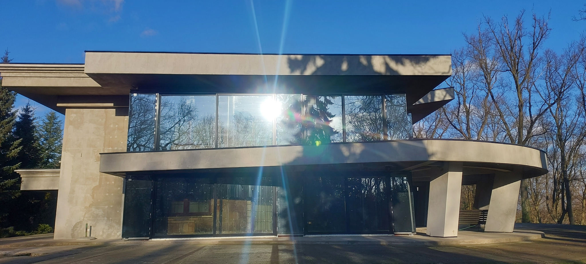 Dwukondygnacyjny budynek z dużymi przeszkleniami, w których odbija się błękitne niebo i korony drzew. Nowoczesna architektura z elementami betonu i naturalnego światła.