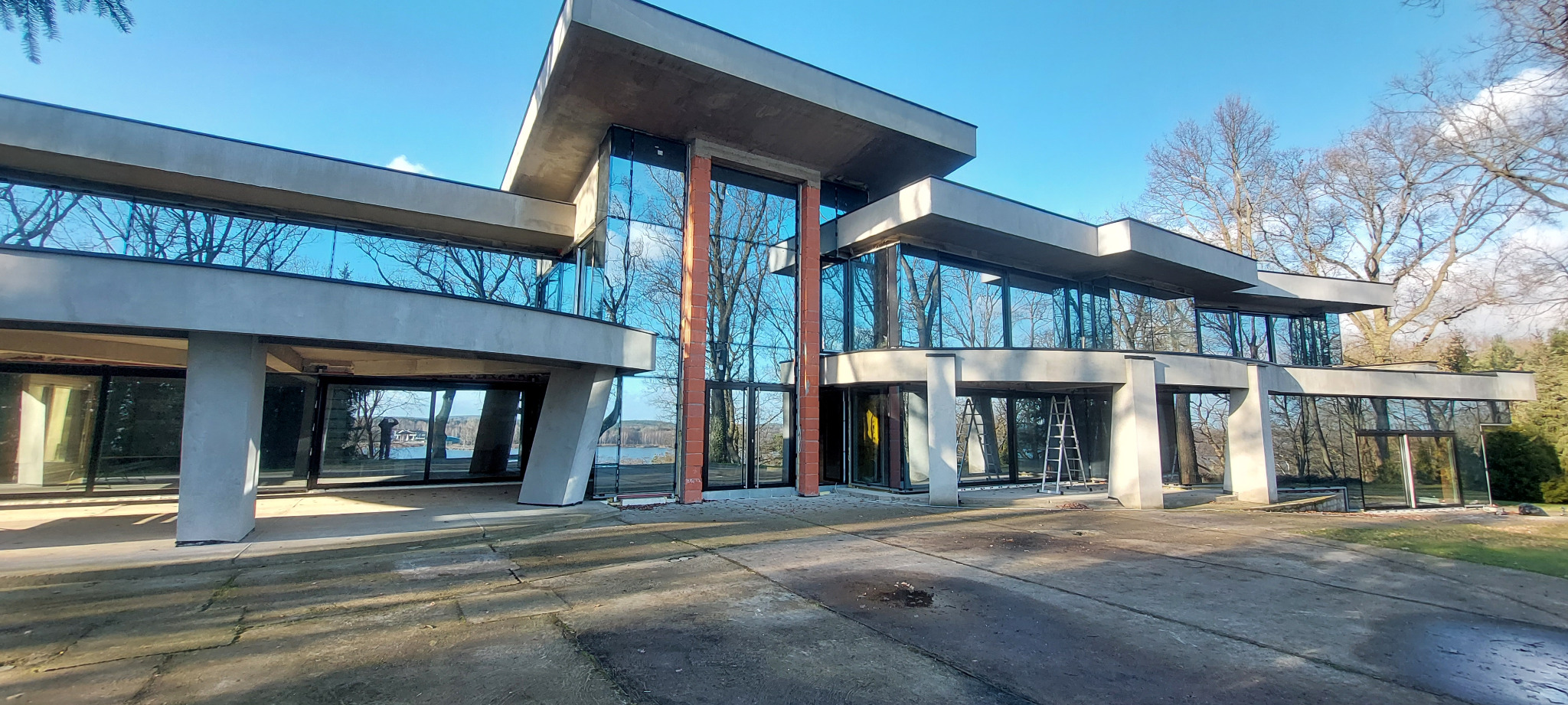 Nowoczesny budynek z dużymi przeszkleniami, częściowo wykończony, z widocznymi elementami konstrukcyjnymi i odbiciem drzew w szybach. Na zewnątrz drabina.