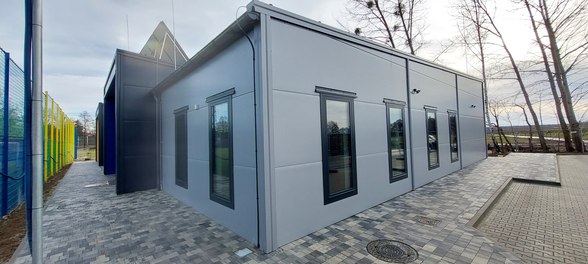 Szara, nowoczesna bryła budynku z ciemnymi ramami okiennymi i kostką brukową. Widok z zewnątrz, w tle drzewa bez liści i fragment kolorowego ogrodzenia.