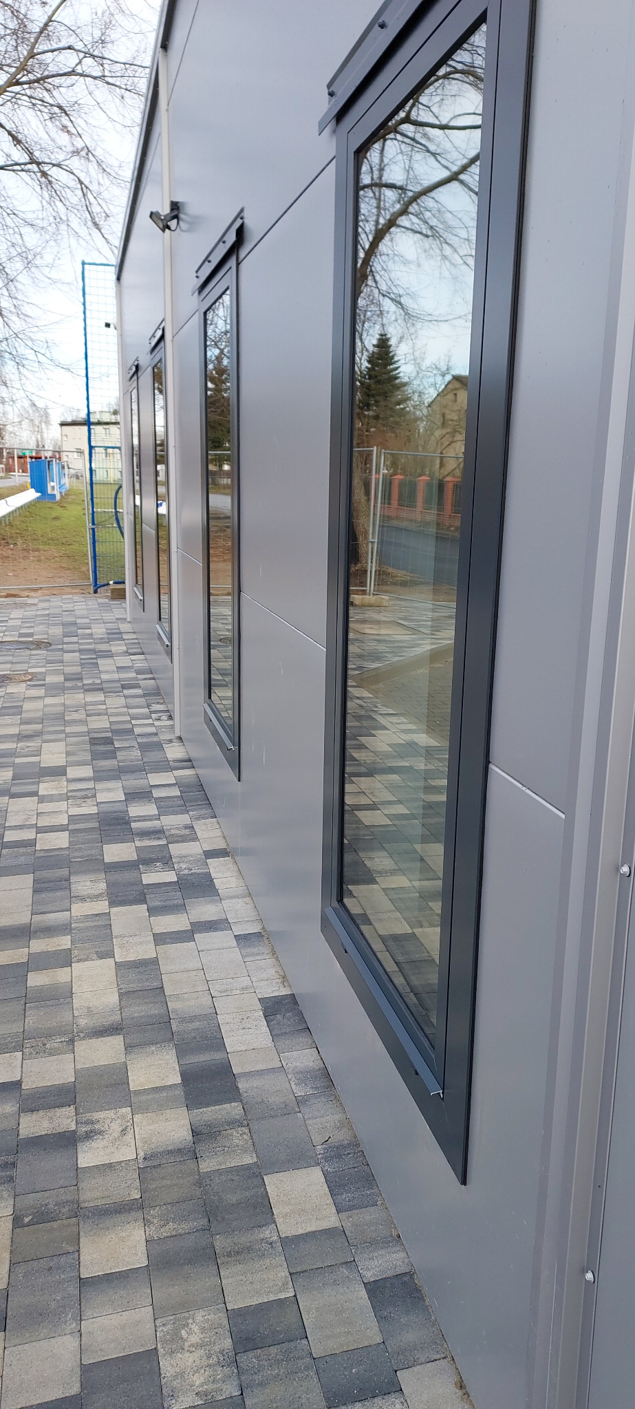 Elewacja budynku z szarymi oknami. W oknach odbija się drzewo. Kostka brukowa w odcieniach szarości. Widoczny fragment boiska w tle, za ogrodzeniem.