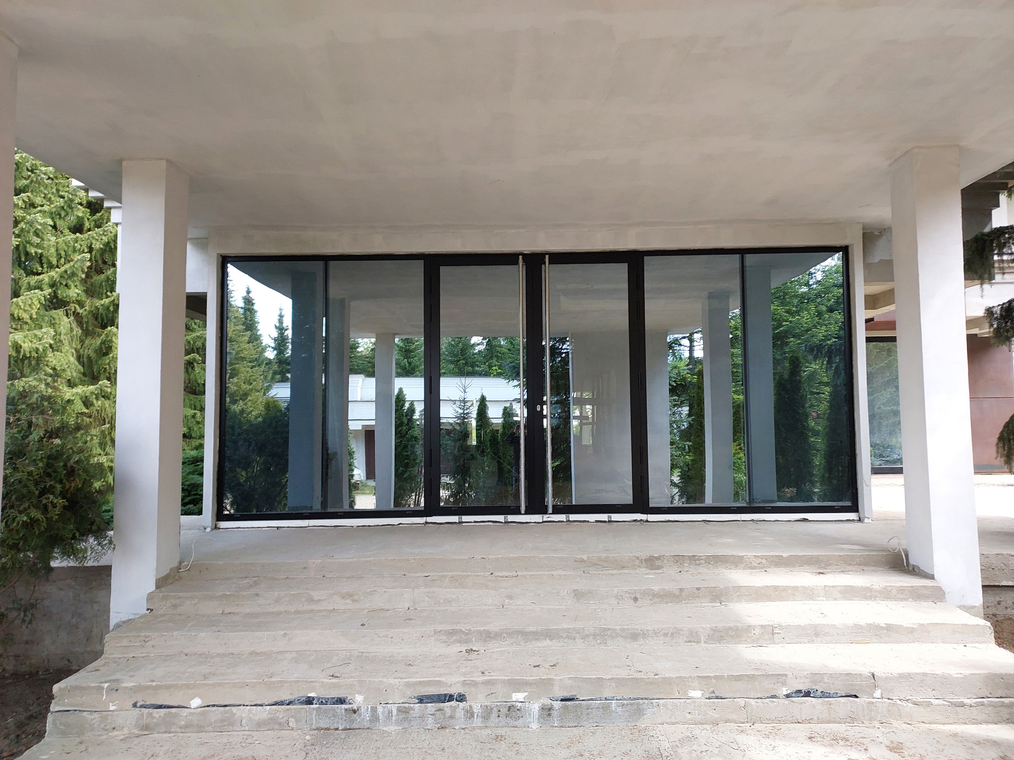 Nowoczesne, czteroskrzydłowe okno aluminiowe z czarnymi ramami, zamontowane w jasnym budynku z kolumnami, widoczne z zewnątrz. Odbicie drzew w szybach.