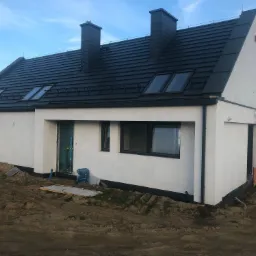 Nowoczesny, parterowy budynek z białymi ścianami i czarnym dachem z lukarnami, widoczna drabina oparta o ścianę, teren wokół budynku w trakcie prac ziemnych.