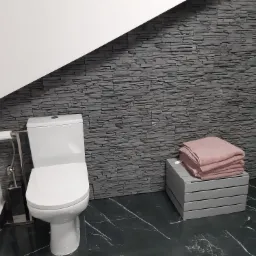 Nowoczesna łazienka z toaletą, szarą ścianą z imitacji kamienia, czarną marmurową podłogą i stosem r&oacute;żowych ręcznik&oacute;w na drewnianym stołku.