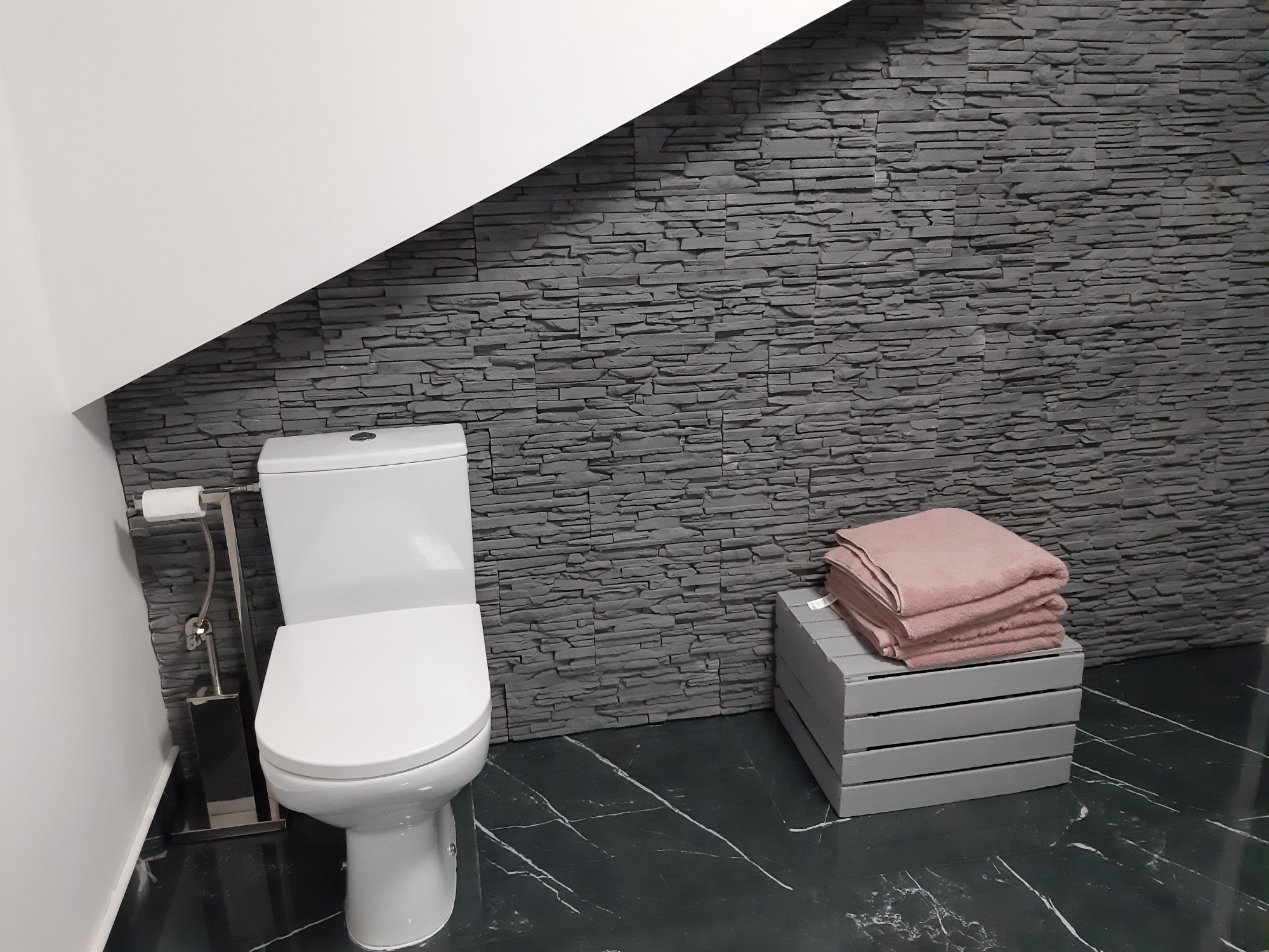 Nowoczesna łazienka z toaletą, szarą ścianą z imitacji kamienia, czarną marmurową podłogą i stosem różowych ręczników na drewnianym stołku.