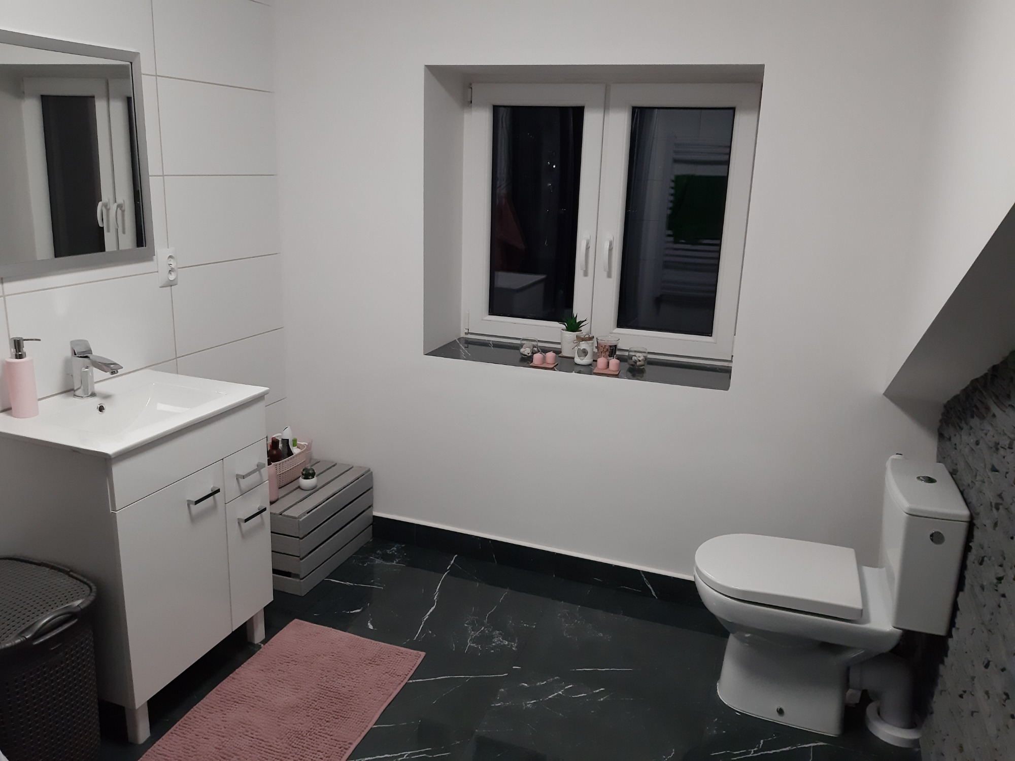 Nowoczesna, jasna łazienka z czarną marmurową podłogą, białymi płytkami, umywalką z szafką, toaletą i oknem z dekoracjami.