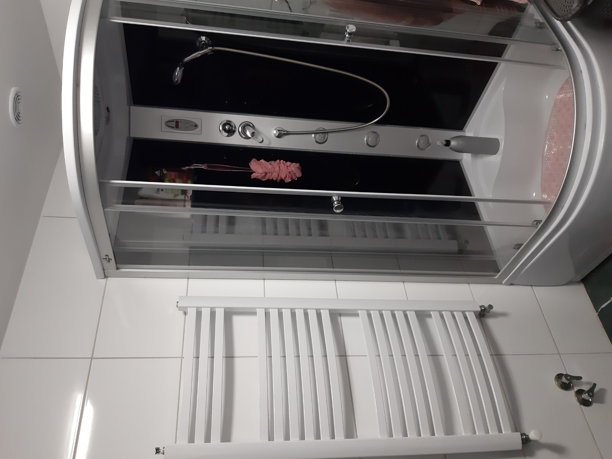 Nowoczesna kabina prysznicowa z hydromasażem, widoczny panel sterowania i deszczownica, obok grzejnik drabinkowy na białych płytkach.