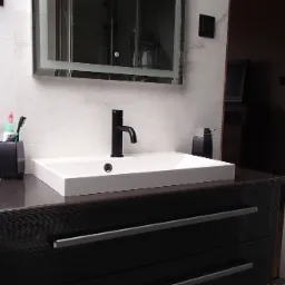 Nowoczesna łazienka z białą umywalką nablatową, czarną baterią i szafką z ciemnego drewna z szufladami na tle marmurowych płytek ściennych.