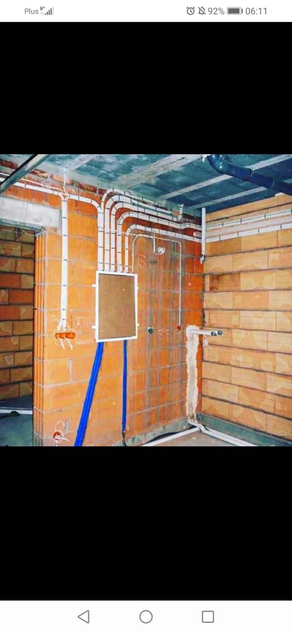 Instalacja elektryczna w surowym wnętrzu: białe rurki prowadzące przewody ułożone na ceglanej ścianie, nieotynkowana ściana z puszkami elektrycznymi i niebieskimi kablami, widoczny strop betonowy.