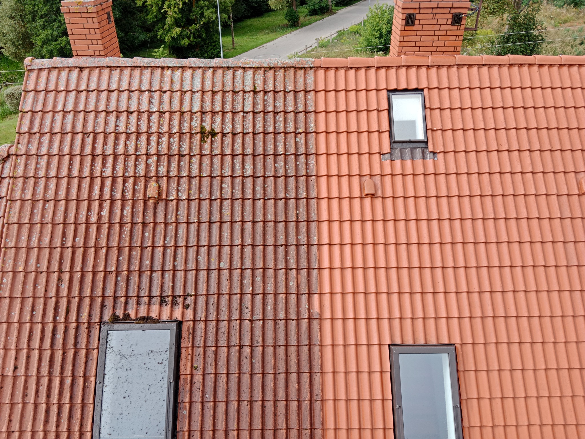 Częściowo wyczyszczony dach z czerwonej dachówki, widoczna wyraźna różnica między brudną i czystą powierzchnią, dwa okna dachowe i ceglany komin w tle.
