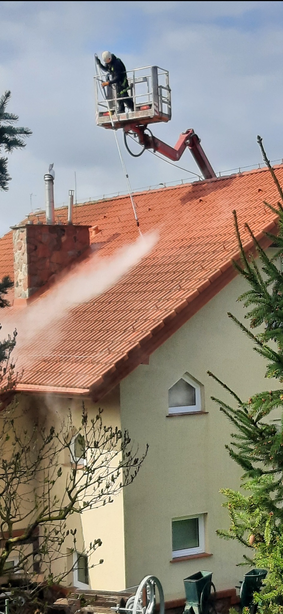 Pracownik w kombinezonie i kasku na podnośniku koszowym czyści dach domu z ceramicznej dachówki za pomocą myjki ciśnieniowej, obok widoczny komin z kamienia. Para wodna unosi się w powietrzu.